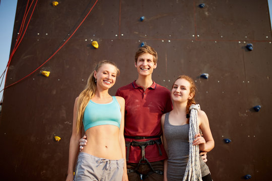 Group of three rock climbers with safety equipment smiling and looking at camera against artificial climbing wall outdoors. Climbing instructor with two girls.