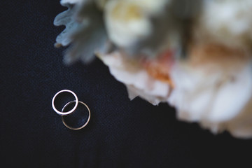 Two gold wedding rings with a diamond on a black background with a wedding bouquet