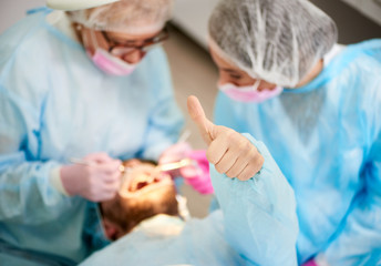 Photo from above doctor woman and her assistant dressed in a one-time uniform work with a male patient who shows a gesture class in a modern dentistry office