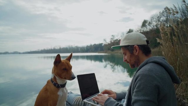 Steadycam slow motion shot of middle aged man, businessman or freelancer work on laptop in park on lake boardwalk, remote office location together with best friend puppy pet