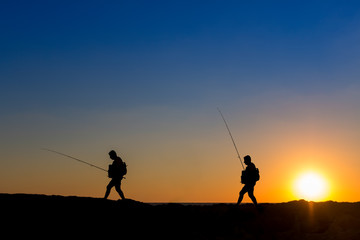 Fototapeta na wymiar 3 Fishermen silhouette with upright rods stroll walk on the rocks at dawn dusk sunrise sunset. The sky is blue purple orange. They have upright rodsat dawn 