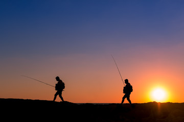 3 Fishermen silhouette with upright rods stroll walk on the rocks at dawn dusk sunrise sunset. The sky is blue purple orange.  They have upright rodsat dawn  
