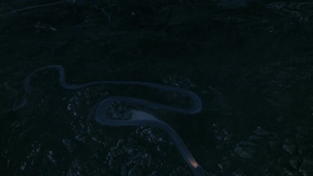 Nighttime drone aerial footage of midnight dark covered dangerous mountain road with turns and switchbacks, car carefully drives through with cautious driver and headlights on. Added film grain style