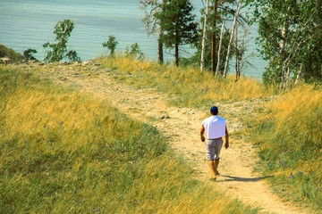 Naklejka na ściany i meble A man in shorts, a white T-shirt and a blue cap walking along a sandy and stony path among bushes, grass and flowers.