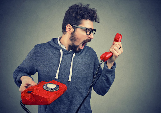 Side view of casual bearded man holding red telephone receiver and yelling in anger. 