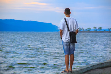 A guy in a white T-shirt and blue shorts stands on the dock on the river bank against the backdrop of the sky and mountains and waits for his love.
