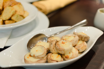 Photo with shallow depth of focus - dish of baked mushrooms champignons with melted cheese in special plate of white color. Serving in restaurant, in background baked potatoes.