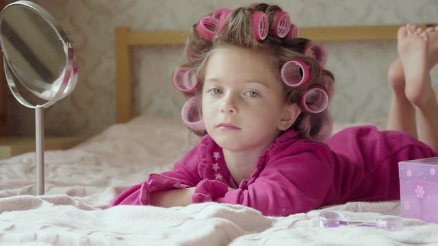 Charming little girl with curlers lies on bed