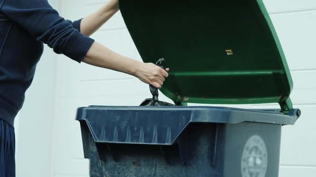An unrecognizable woman throws a bag of garbage into a garbage can near the house