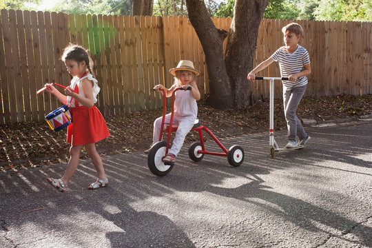 Three children in mini parade, banging drum, riding tricycle and using scooter