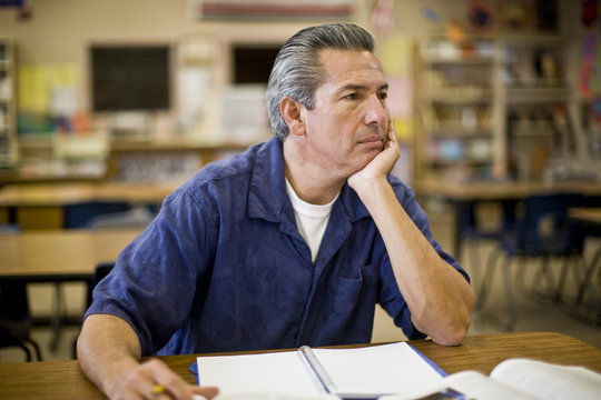 Mature man sitting at a desk in a classroom with his chin in his hand.