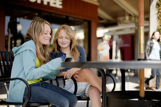 Two teenage girls looking at a cell phone and smiling while at a cafe