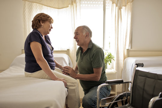 Mature female patient sitting on hospital bed, her husband kneeling beside her holding her hand.