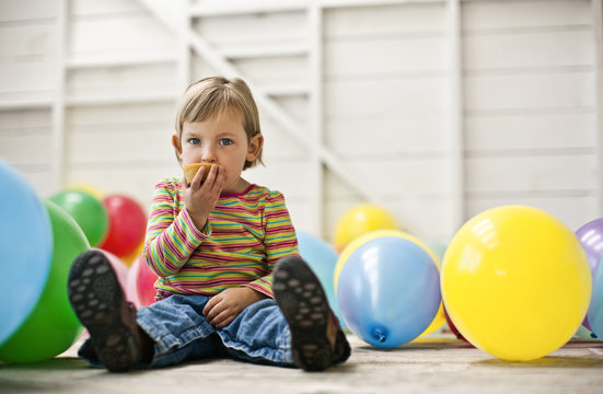 Little girl sitting in a room full of balloons and eating a cupcake.