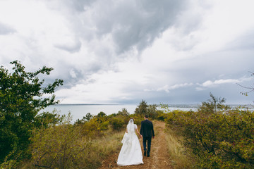 Beautiful wedding photosession. Handsome groom in blue formal suit and bow tie with boutonniere and his elegant bride in white dress and veil with a beautiful hairdress on a walk in field near river