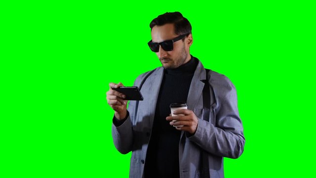 Man in suit and black sunglasses use mobile and hold coffee cup. Green screen, footage.