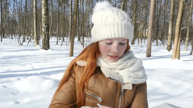 Red-haired girl staying at winter park and draws in sketchbook. Beautiful teenage girl painting on paper