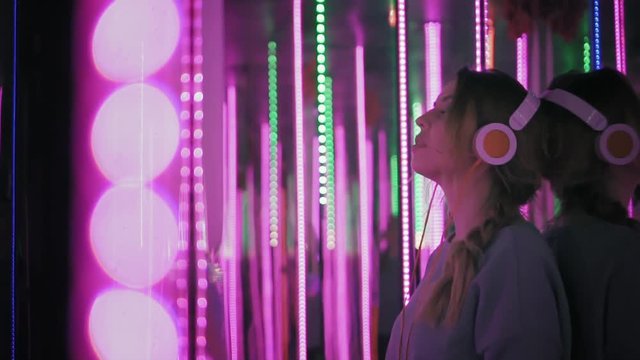 Hipster female in earphones leans to the mirror, looking in camera and smiling. Woman listen music in neon lights at party. Slow mo