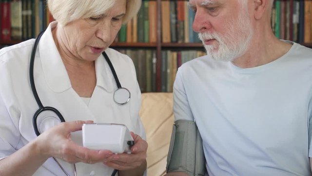 Female professional doctor in white coat with stethoscope at work. Senior woman physician measuring pressure to sick senior male patient by tonometer at home. Consulting about treatment and therapy