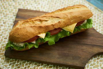 Fresh submarine sandwiches with chicken breast cheese, tomatoes, lettuce, cucumbers and onions on dark wooden cutting board, rustic background
