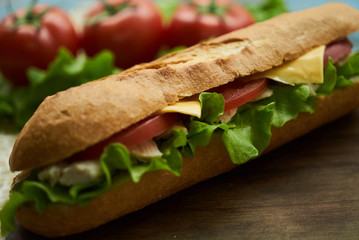 Baguette sandwich with turkey breast, cheese, lettuce, tomatoes and onion on a cutting board. Long subway sandwich on a rustic background, close-up