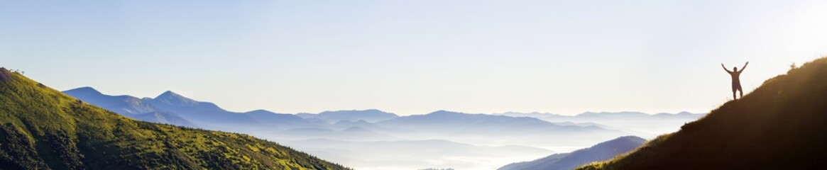 Panorama of young successful man hiker silhouette open arms on mountain peak.