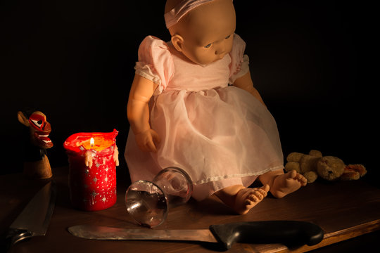 Terror thriller concept baby doll scene with assassin murder knives, spilled wine glass, spooky gloomy candle and devil figure on dark background