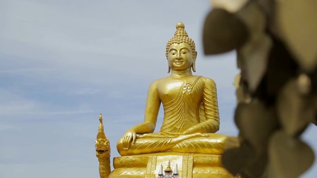 Huge golden sculpture of Buddha in Tailand. Buddhist God is sitting in traditional cross-leged pose, with the hands og his knees. Cloudy light sky are at the background of religious image and bronze