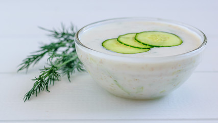 Tzatziki in a glass bowl with cucumber slices and dill on a white wooden table.