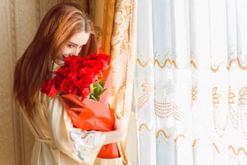 Nice romantic sweet woman with bouquet of red roses in beige interior apartment. Lady in silk robe at cozy home. Concept of tenderness and woman's feeling 