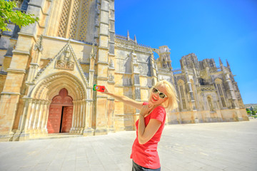 Fototapeta premium Happy woman takes photo of Batalha Monastery one of the most important Gothic sites in Portugal. Female lifestyle takes pictures by mobile phone with Portugal flag cover.Tourism and travel in Portugal
