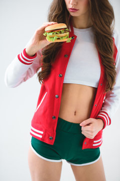 cropped image of seductive girl in baseball jacket holding burger isolated on white