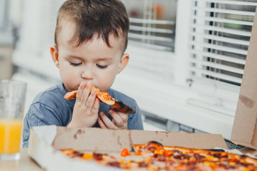 the little boy eats a huge harmful pizza himself in the kitchen and drinks juice, very fat and harmful