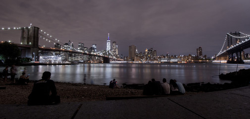 On a summer evening local people and tourists on Brooklyn’s Pebble Beach foreshore take in New York’s magnificent cityscape brightly lit at night framed by the stunning Brooklyn and Manhattan Bridges