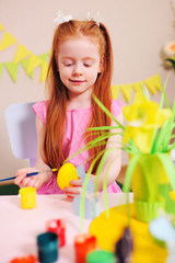 cute little girl with red hair painting a yellow eggs on the background of the Easter decor