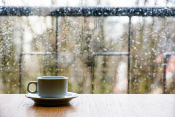 A white espresso cup on a wooden table in front of a glass window covered with rain drops.