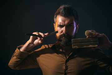 Professional barber. Silhouette bearded man, macho with beard and moustache, holds in hands straight razor, wooden casket, shaving brush. Bearded hairdresser in stylish clothes holds straight razor.