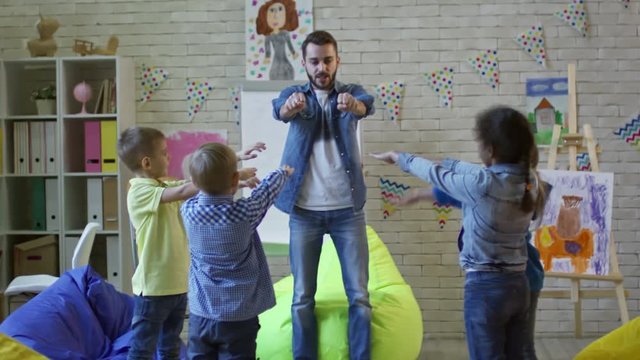 Young male teacher and group of little kids doing wrist exercises and raising arms during kindergarten lesson