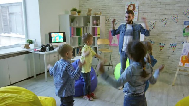Young male teacher talking and doing shoulder exercises with group of little cute kids in kindergarten classroom