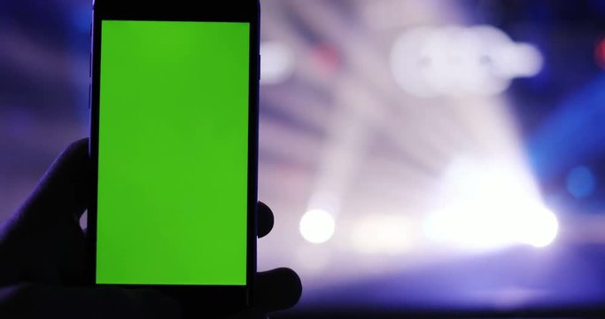 closeup view of modern smartphone on blurred colorful cocert lighting background / man hand holding mobile green screen chromakey mock-up photo camera night club music clubbing app stream chat wifi