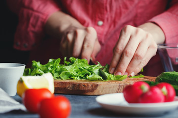 Girl in a pink shirt slices a fresh salad on a textured wooden board surrounded by vegetables. A series of photos about the preparation of summer vegetable salad.