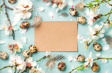 Easter holiday background. Flat-lay of tender Spring almond blossom flowers on branches, feathers, quail eggs and paper in center over blue background, top view, copy space. Greeting card concept