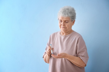 Elderly woman with diabetes measuring level of blood sugar on color background