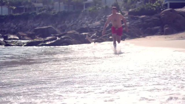 A slender man in red shorts runs along the sea waves at the shore, splashes scatter in different directions. slow motion, 1920x1080, full hd