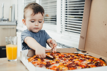 the little boy eats a huge harmful pizza himself in the kitchen and drinks juice, very fat and harmful