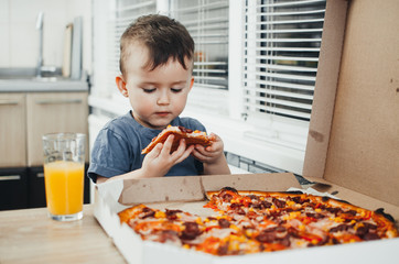 the little boy eats a huge harmful pizza himself in the kitchen and drinks juice, very fat and harmful