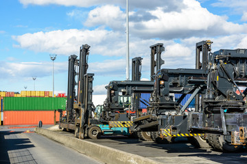 A row of reach stackers and carrier trucks parked on the container storage platform of the intermodal terminal of a river port with containers in the background.