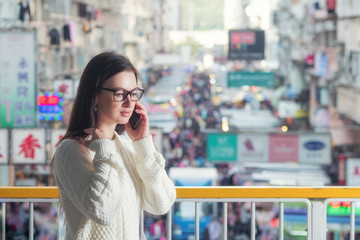 Young attractive caucasian brunette woman in eyeglasses standing on pedestrian footbridge and talking mobile phone. Blurred traditional Hong Kong street market as background. 