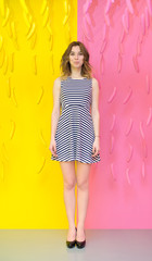 beautiful girl standing and smiling against a background of 2 colors dividing background in half a pink and yellow, saturated and weaving in the air painted bananas. Striped dress and evening shoes.