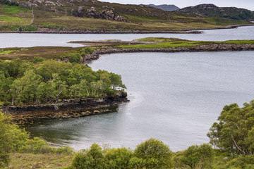 Balgy, Scotland - June 10, 2012: Closeup of peninsula in Silver colored Upper Loch Torriden. Green hills with trees in front. Mountains on horizon.